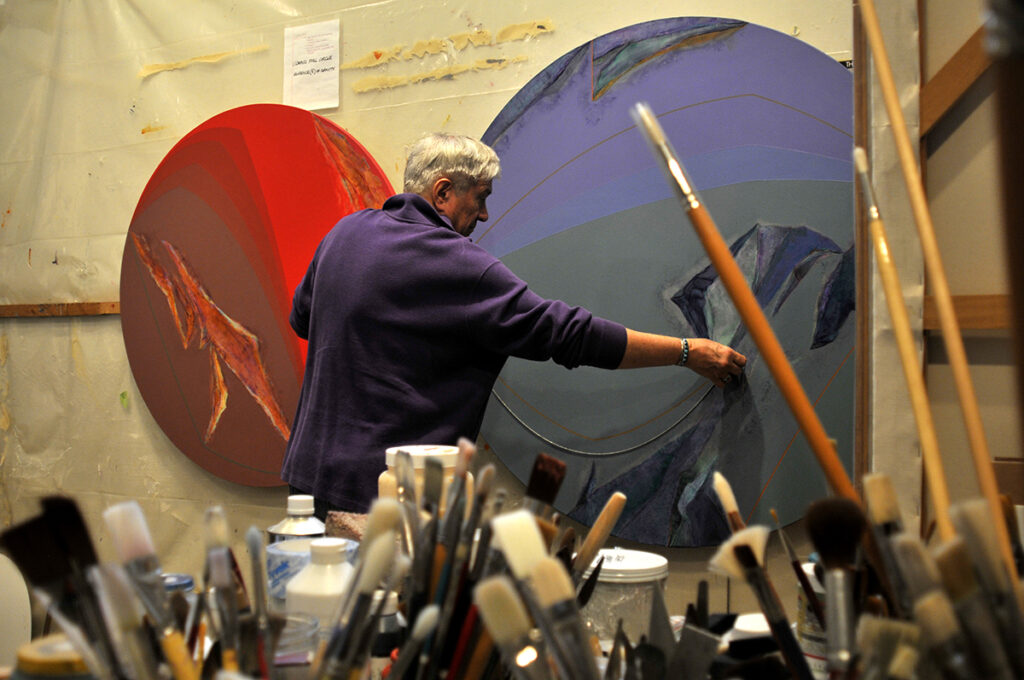 A white-haired man wearing purple holds a chain up to an abstract painting to demonstrate a catenary curve in the work.